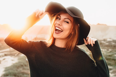Young woman in glasses and holding hat and winking on the beach with sunset light